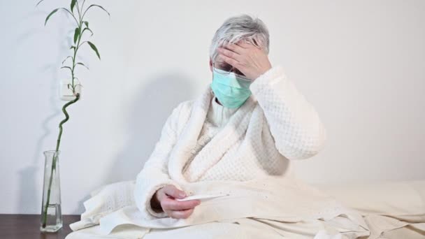 Coronavirus. Malade femme âgée avec masque de protection tenant thermomètre assis sur le lit, contrarié vieille femme mûre attrapé froid eu coronavirus symptômes de grippe à la maison seul . — Video