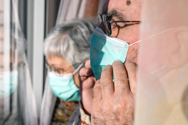 Para seniorów, w maskach ochronnych, w domu patrząca przez okno. Koncepcja kwarantanny dla koronawirusów zostaje w domu i oddala się od społeczeństwa. Zamknięci ludzie. w podeszłym wieku i na emeryturze. — Zdjęcie stockowe