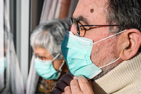 Starší pár s ochrannými obličejovými maskami, doma, dívající se oknem. Koncept koronavirové karantény zůstat doma a sociální distancování. Uzamknout lidi. starší osoby a důchodový životní styl. — Stock fotografie