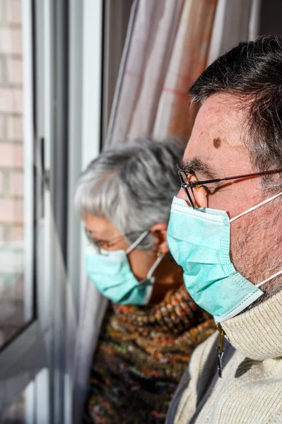 senior couple, with protective face masks, at home looking through the window. Concept of coronavirus quarantine stay home and social distancing. Lockdown people. elderly and retired lifestyle.