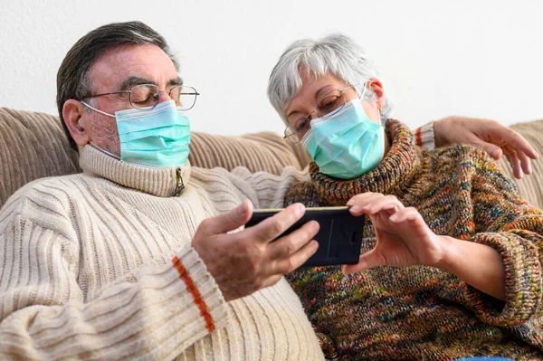 Coronavirus. Stay at home, lifestyle. Cheerful Elderly couple sitting on a sofa on a quarantine at home, making a video call with the smartphone. Senior Couple wearing protective masks.
