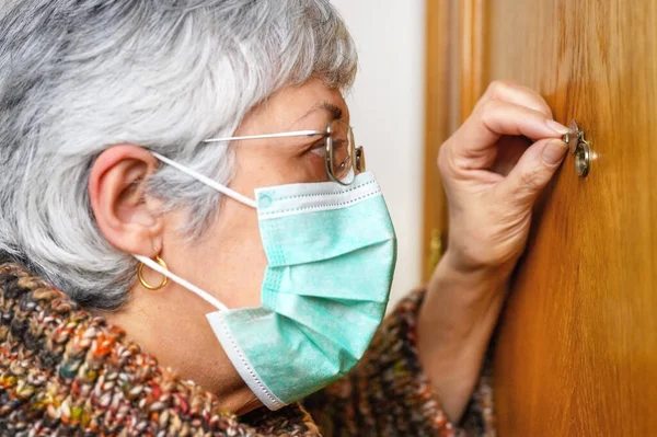 Covid-19 concept. Stay at home. Self-isolation to prevent the coronavirus pandemic. Senior woman in protective green mask looks through the peephole. — Stock Photo, Image