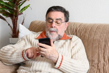 Coronavirus karantinası sırasında evdeki koltukta oturan yaşlı adam cep telefonu kullanıyor..