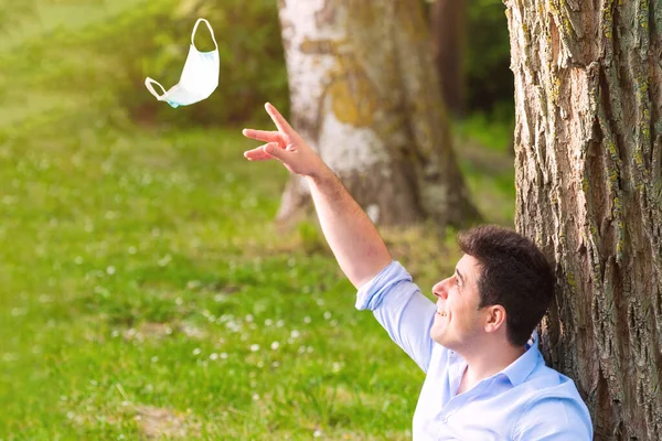 Young man sitting in the park throwing away protective face mask celebrating victory over coronavirus outside in open air. End of quarantine. Remove mask. Outdoor. Freedom. Pandemic crisis end. — Stock Photo, Image