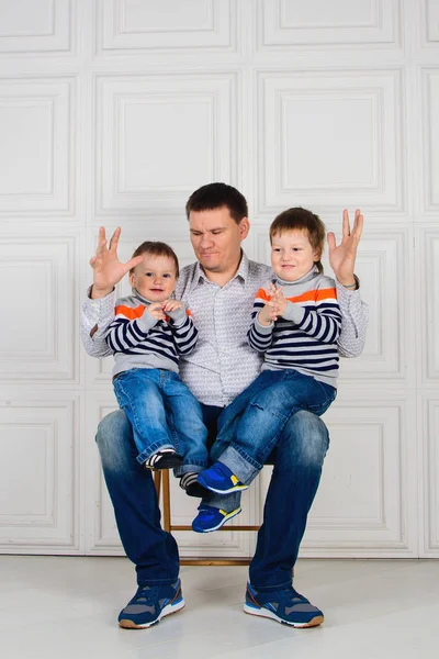 Dad with two children on his knees sitting on the chair in front of white wall. Children in identical sweaters — Stock Photo, Image