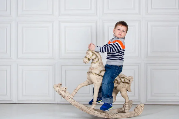 Un ragazzino che dondola su un cavallo di legno. serio ragazzo di tre anni in jeans e maglione su sfondo bianco . — Foto Stock
