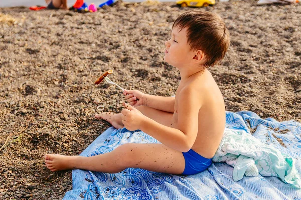 El niño come salchichas fritas. Bronceado? hild en bañadores sentados en la playa — Foto de Stock
