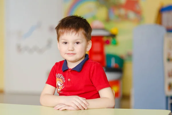 Мальчик в красной футболке сидит за столом в детском саду — стоковое фото