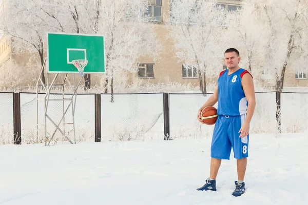 Un homme joueur de basket-ball en uniforme de sport bleu se tient sur un terrain de basket-ball et tient un basket-ball dans ses mains en hiver — Photo
