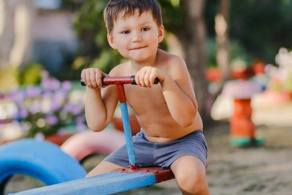Маленький милий хлопчик без сорочки грає на дитячому майданчику, катається на дерев'яних гойдалках — стокове фото