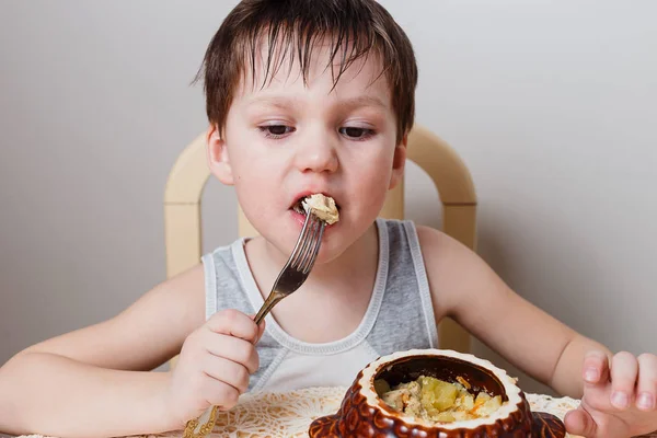 Um menino come frango assado com batatas de uma panela de barro — Fotografia de Stock