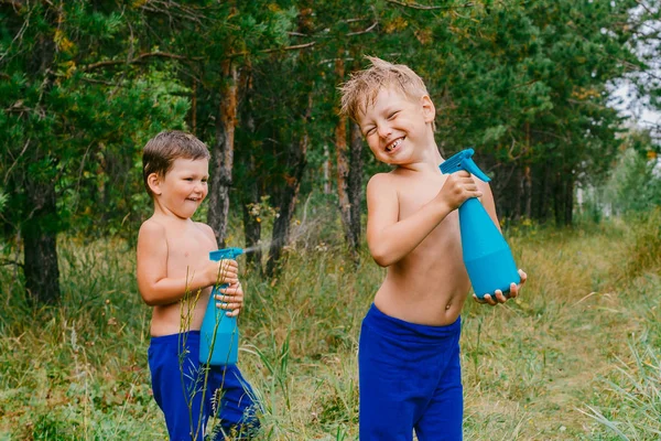 Two cheerful boys in blue pants splash water from sprayers and laugh in the green forest — Stock Photo, Image