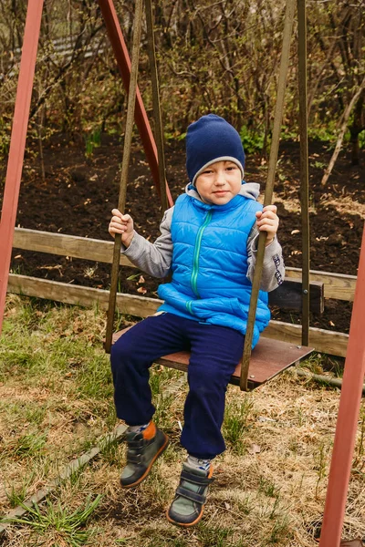 the boy in the blue vest and hat swinging on the iron swing in the garden