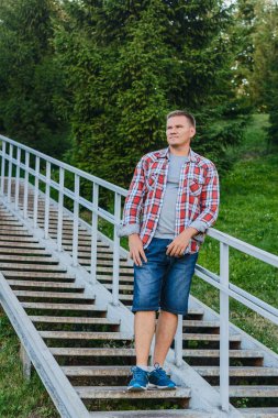 Genç bir adam bir ekose gömlek ve kot şort bir metal üzerinde duruyor 
