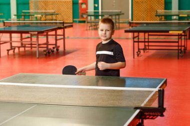 Sekiz yaşında beyaz bir çocuk masa tenisi raketi tutuyor. Çocuklar içeride masa tenisi oynuyorlar.
