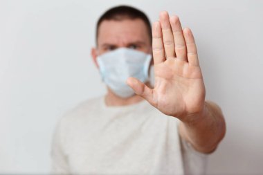 Yüzünde tıbbi bir maske olan orta yaşlı, vahşi bir adam. Avucu beyaz bir arka planda uzanıyor. Koronavirüsü durdur. Evde kal.