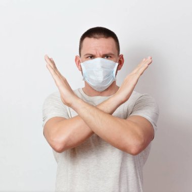 Kollarını beyaz bir arka planda çapraz yapmış tıbbi maskeli ciddi bir adam, koronavirüsün yayılmasını önlüyor.