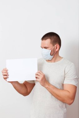 Hafif giysili ve yüzünde tıbbi maske olan ciddi bir beyaz adam beyaz arka planda boş bir sayfa tutuyor. Kağıda bak. Evde kal.
