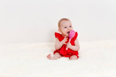 Sekiz aylık kırmızı elbiseli bir kız bebek evde bir yatakta otururken pembe bir çıngırağı kemiriyor.