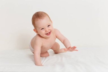 8 aylık gülümseyen bir bebek beyaz bir çarşafın üzerinde emekliyor. Mutlu, çıplak bir bebek.