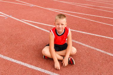 Stadyumdaki koşu bandında bir çocuk oturuyor. Koşudan sonra çocuk