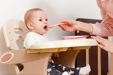 Bebek ilk yemi yemek için bir çocuğun sandalyesinde oturarak yer. Bebek ağzını açar.