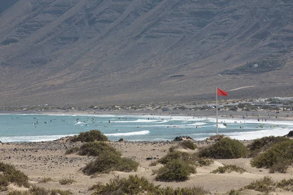 Rudá vlajka váží ve větru na pláži Famara surfaři na Lanza — Stock fotografie