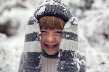 Karla kaplı sıcak eldivenler içindeki bir çocuk iki eliyle yanaklarını tutar ve gülümser.