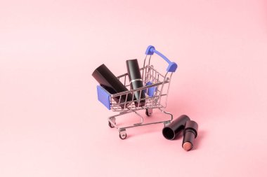 Siyah kutulardaki rujlar parlak pembe bir arka plandaki alışveriş arabasında bulunur. Kozmetik alım konsepti, online alışveriş