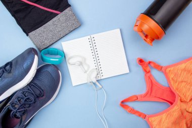 Spor sahasının düz düzeni. Spor ayakkabıları, kulaklıklar, kıyafetler, şişe, saat ve mavi arka planda yazmak için açık bir defter. Spor Planlama.