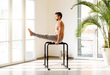 Fit muscular man doing V sit calisthenics exercises on parallel bars to tone and strengthen his muscles in a bright airy gym with copy space in a health and fitness concept clipart