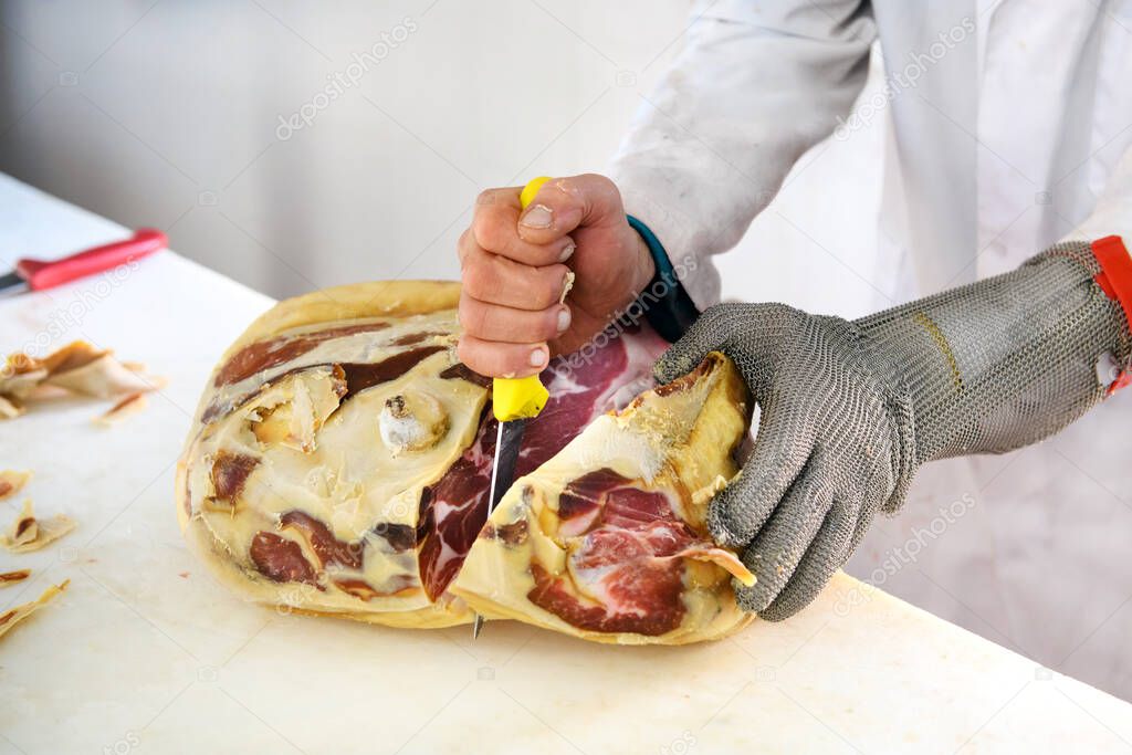 Butcher with short sharp knife deboning pice of prosciutto ham meat, using wire mesh cut resistant glove