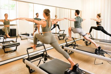 Yenilikçi yataklar üzerinde hareketli hamleler yapan pilates sınıfı sporcuları, sağlık ve zindelik konseptinde yüksek anahtarlı bir spor salonunun aynasına yansıtılıyor.