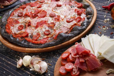 Siyah hamurdan yapılmış pizza, ahşap bir masada et ve sosis. Pizza tahtasındaki malzemeler..