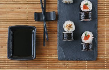 Suşi, somon, ton balığı, avokado, alglerde pirinç ve bambu hasırının üzerinde duran siyah bir tahtanın üzerinde çubuk. Japon, Asya yemekleri...