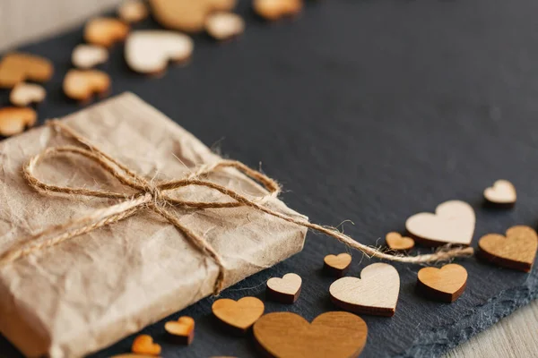 Mały prezent zawinięty w papier kraft. Małe drewniane serca na tle. — Zdjęcie stockowe