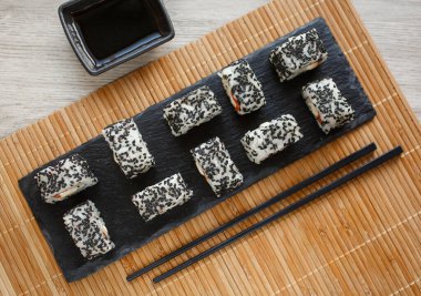 Minimalizm ve sofistike: lezzetli somon balığı ve siyah susam tohumlarıyla kaplı avokado suşisi. Siyah bir taşın üzerinde zencefil ve wasabi ile servis edilmiş. Üst görünüm.