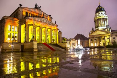 Ünlü Gendarmenmarkt ışıklı Berlin konser salonu ve Fransız Katedrali ıslak zemin üzerinde yağmur sonrası alacakaranlık günbatımı sırasında Berlin şehir merkezinde yansıtan kare panoramik manzaralı