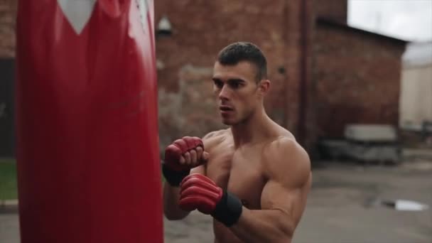 Vista lateral cercana del boxeador masculino muscular practicando golpes y patadas con un saco de boxeo al aire libre. Pared marrón en el fondo — Vídeo de stock