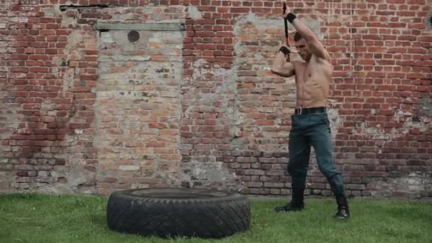 Strong muscular shirtless young man hitting a tire with a sledgehammer. Red brick building on the background — Stock Video