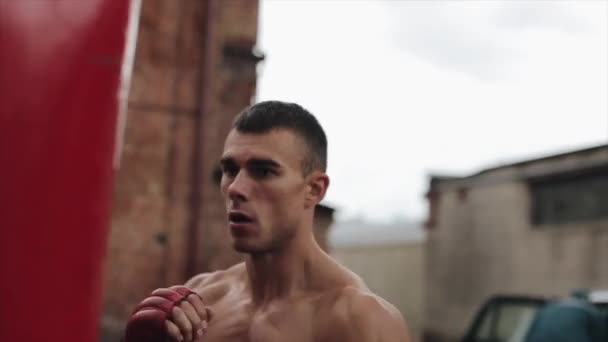 Bliski widok z boku muskularny bokser męski ćwiczenia uderzeń i kopnięć z workiem bokserskim na zewnątrz — Wideo stockowe