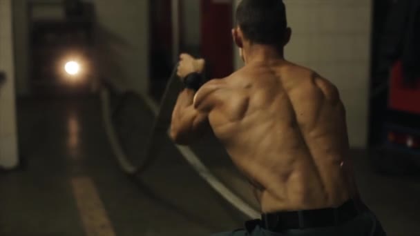 Тесный вид сзади на мускулистого пожарного, который тренируется, держа два развернутых пожарных шланга и быстро двигая руками — стоковое видео