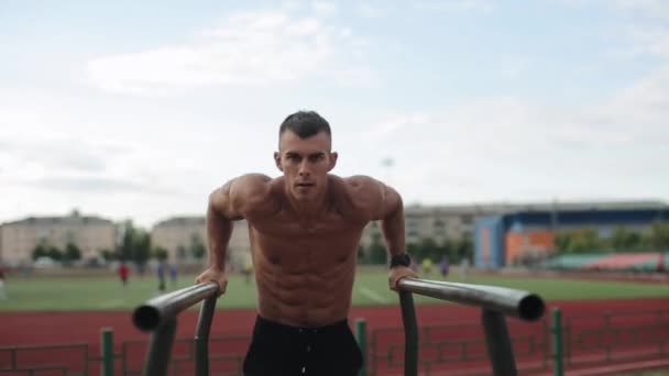 Close view of a shirtless muscular athlete doing bodyweight push-up working out on the sports ground. Blurred view of people playing football on the background — ストック動画