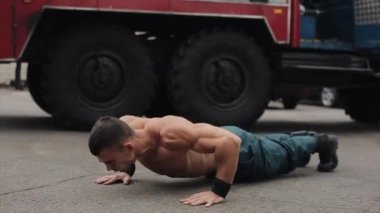 Ağır çekim. Çekici, üstsüz, zıplayarak şınav çeken genç bir sporcu. Arka planda büyük bir itfaiye arabası var. Yan görünüm