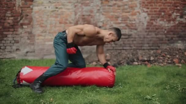 肌肉男运动员坐在户外的长沙袋上，拳打脚踢，倒立，不停地打沙袋 — 图库视频影像