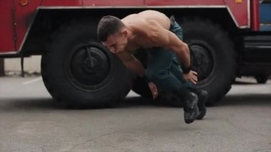 Ağır çekim. Çekici, üstsüz, zıplayarak şınav çeken genç bir sporcu. Arka planda büyük bir itfaiye arabası var. Yan görünüm