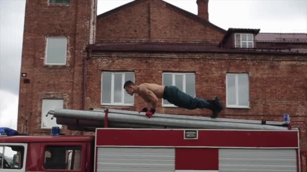 Fuerte bombero sin camisa haciendo flexiones con las piernas cruzadas en la parte superior del camión de bomberos — Vídeos de Stock