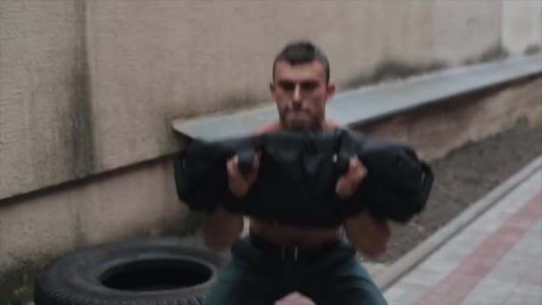 Jóképű félmeztelen fiatalember guggol és bicepsz gyakorlatokat végez egy nehéz táskával. A szabadban végzett munka — Stock videók
