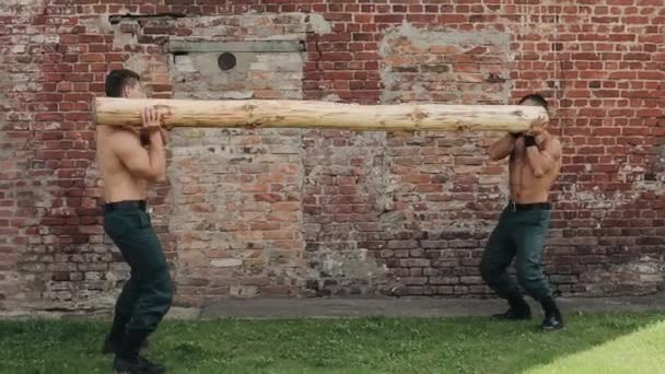 Two young handsome men training outdoors raising a log over their heads. Red brick wall in the background — Stock Video