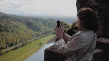 Dresden, Almanya - 12 Ağustos 2019: Güzel genç kız akıllı telefonuyla güzel dağların fotoğraflarını çekiyor. Yan görünümü kapat. Yükseklerde, aşağıda dağ nehri.